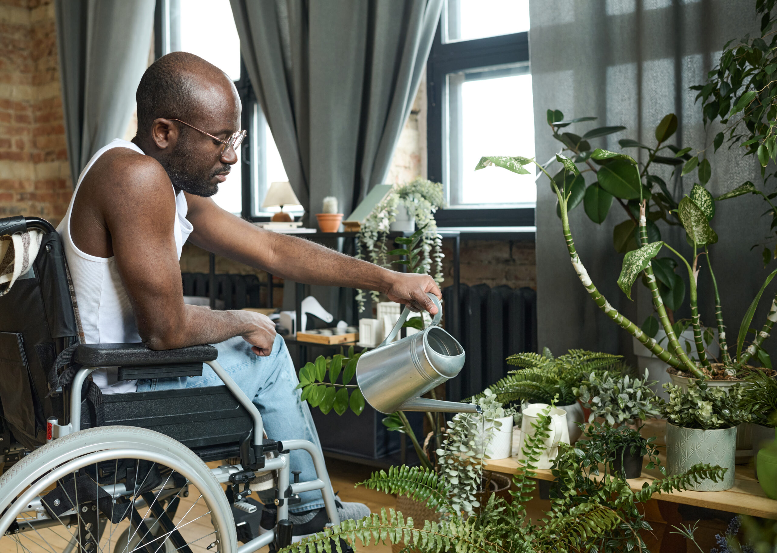 African man in wheelchair watering potted flowers with watering can while having rehabilitation at home