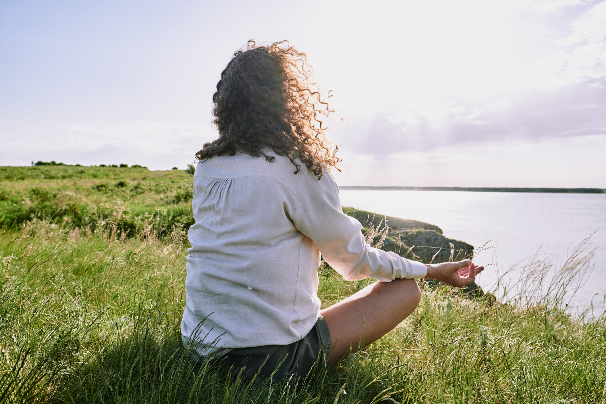 Rear view of curly-haired woman sitting with crossed legs on grass and meditating in silence by tranquil lake - trauma triggers