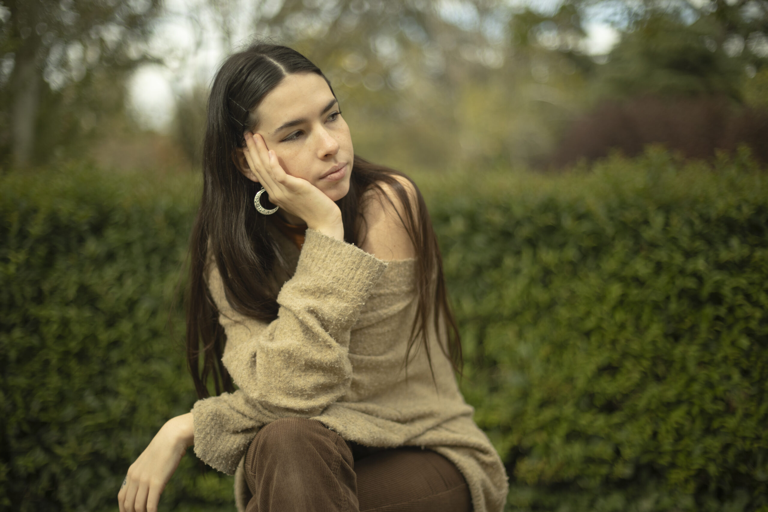 A young Caucasian woman sits outdoors, chin resting on hand against a blurred plant background, directing her gaze towards copy space. versatile visual with emphasis on nature and customizable content - Calming the mind