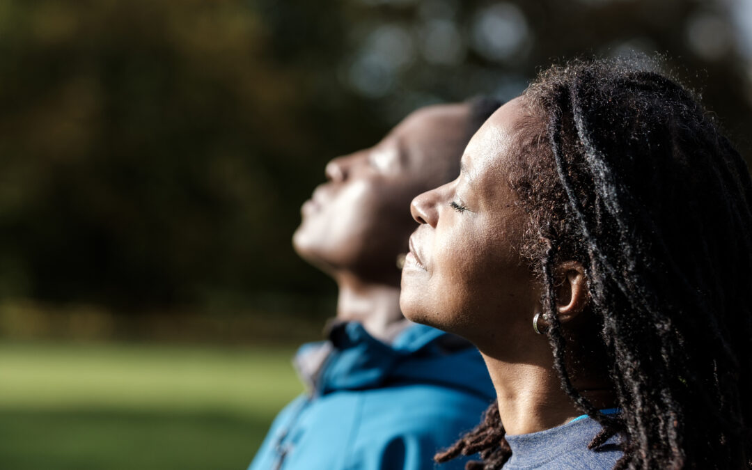 Portrait of two mature black women enjoying sun in an autumn day. They are in a park and they look really relaxed and calm. Their eyes are closed and they are wearing warm sport outfits.